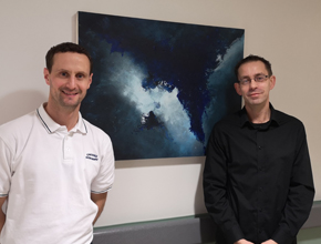 Two people stand for a photo in front of artwork hanging on the wall